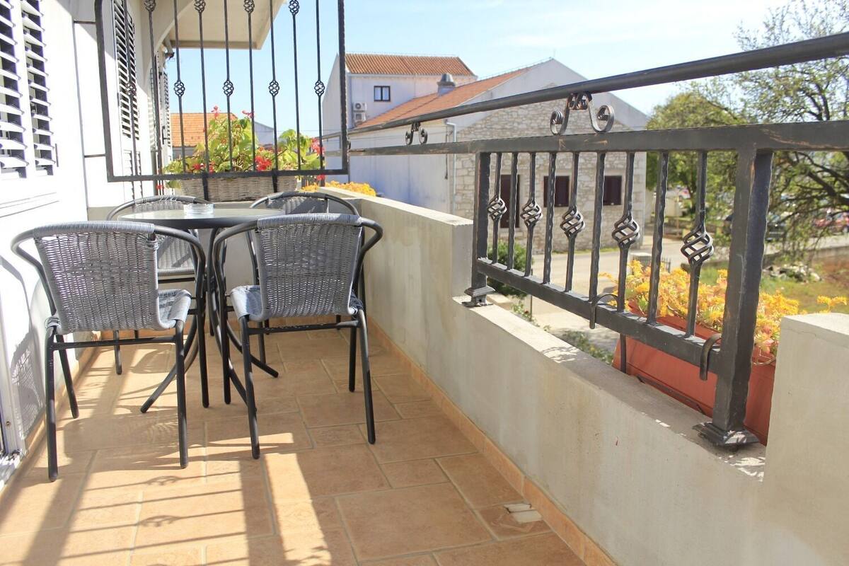 Un balcone con due poltrone in vimini, un tavolino, ringhiera in metallo e vista su una zona residenziale con case e alberi.