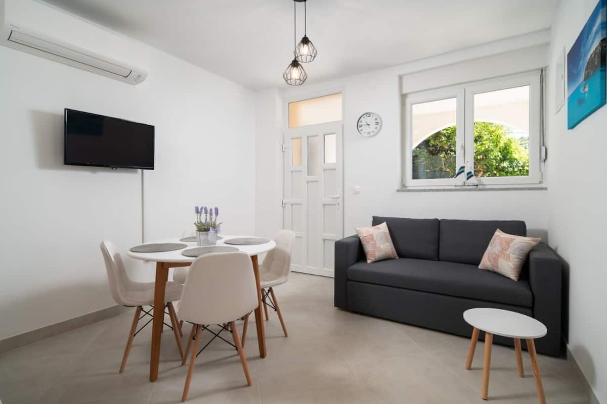 A modern living room with a grey sofa, a small round table with chairs, a flat-screen TV mounted on the wall, and a window with a view of greenery.