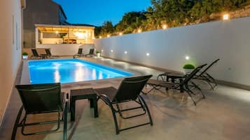 An outdoor swimming pool with lounge chairs and a table, surrounded by a white wall and trees.