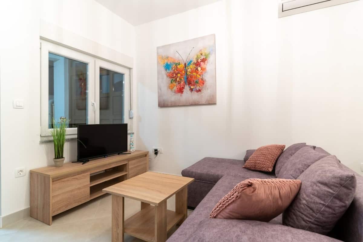 A modern living room with a flat-screen TV on a wooden stand, a grey sofa with a woven throw pillow, and a colorful abstract painting on the wall.