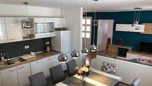 A modern kitchen with white cabinetry, a dining area with a table and chairs, and a living area with a sofa and a TV. - House Franny (Zadar)