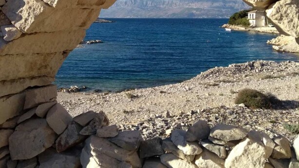 Ein steinerner Bogen führt zu einem felsigen Strand mit freiem Blick auf das Meer und entfernte Berge.