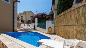 A swimming pool with white lounge chairs, a stone border, and a bamboo fence. - Apartments Dalis (Milna)