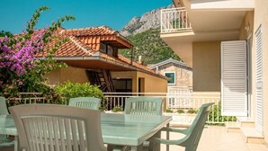 A balcony with a table and chairs, a view of a mountain, and a building with a tiled roof. - Apartments Andelko (Baska Voda)