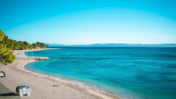 A beach with clear turquoise waters, a sandy shore, and a row of trees. - Apartments Andelko (Baska Voda)