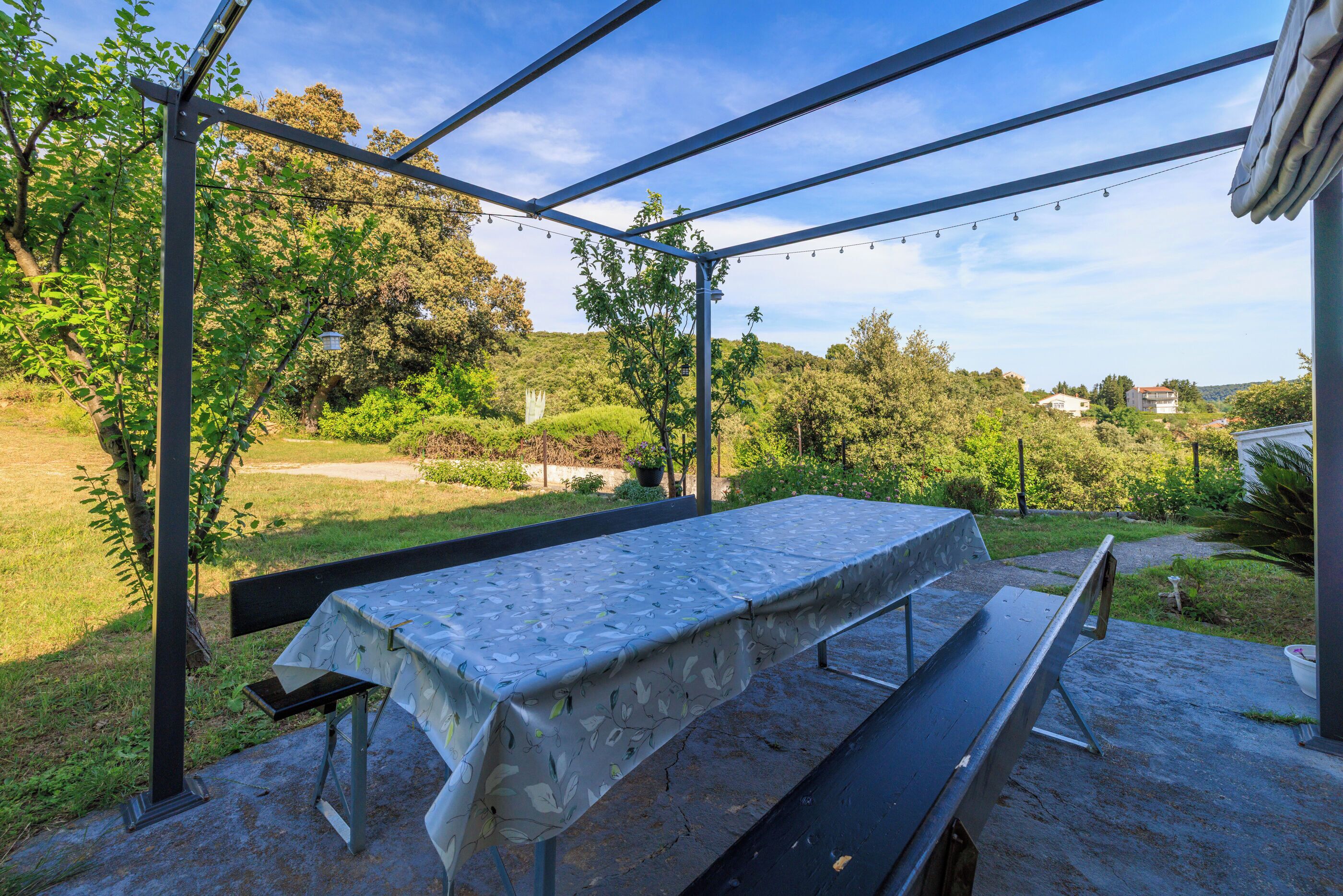 Um pátio externo com mesa e banco cobertos, rodeado por vegetação e vista para o campo.