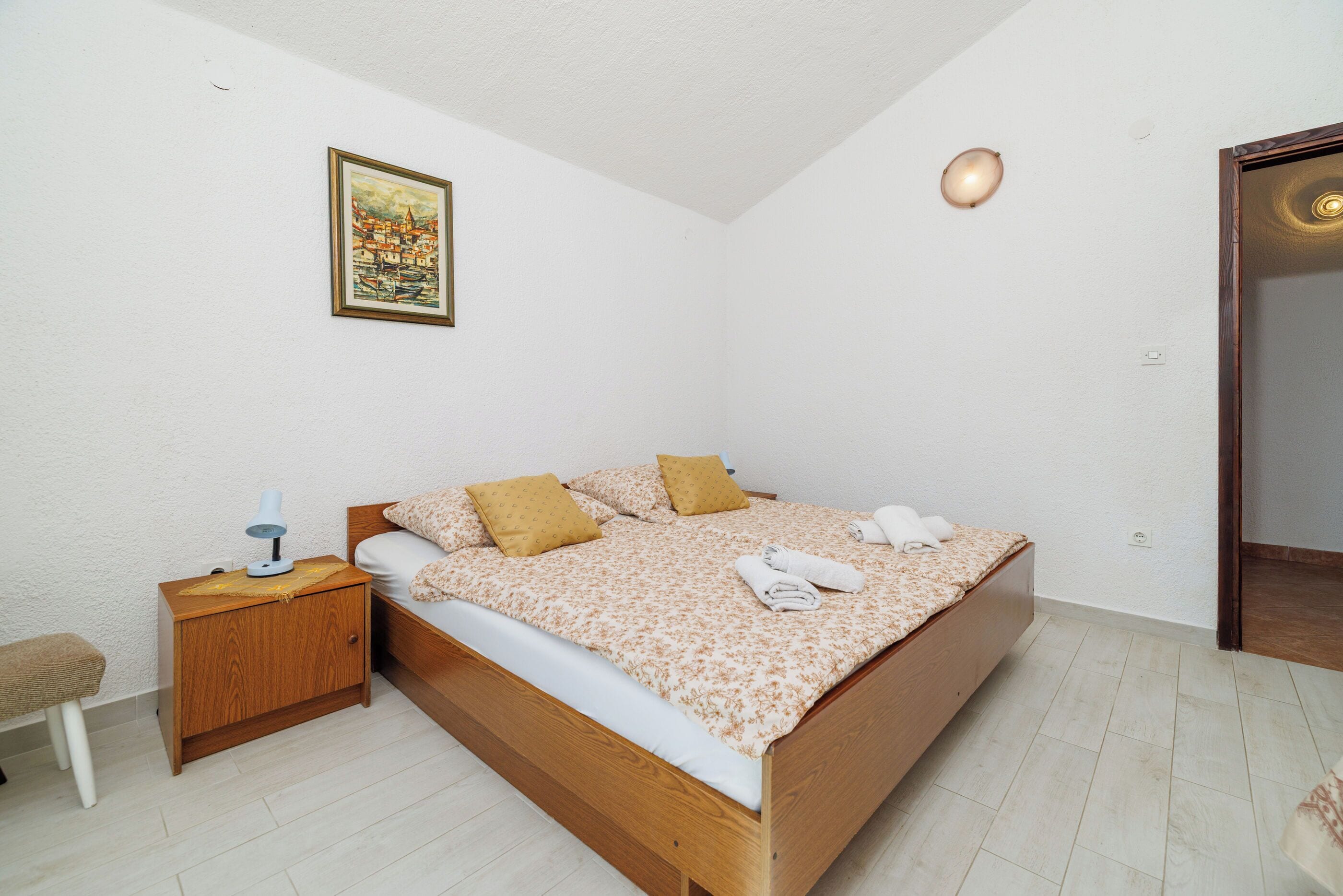 A bedroom with a wooden bed, a nightstand with a lamp, a small stool, and a painting on the wall.