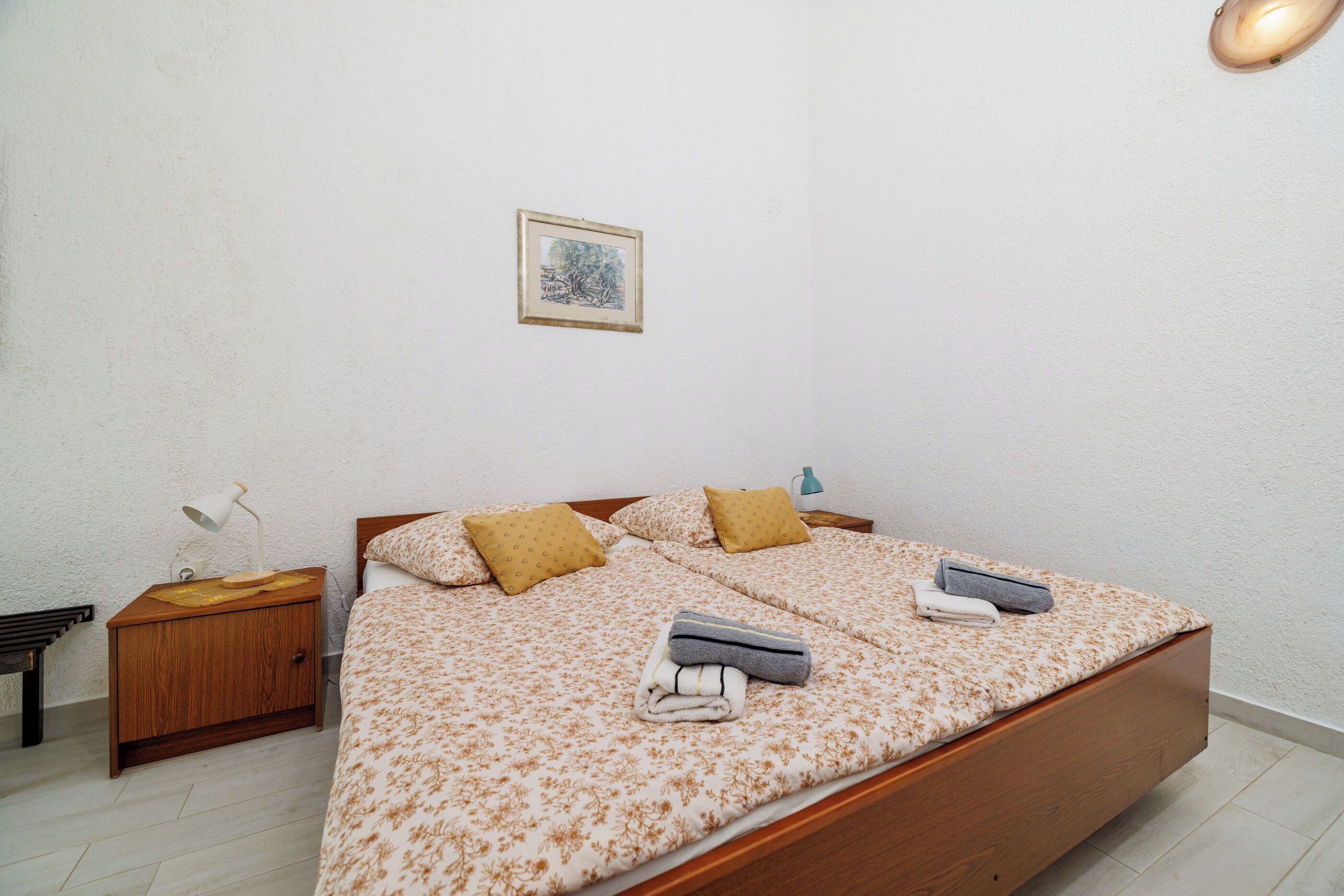 Una cama bien tendida con una colcha estampada con motivos florales, dos almohadas y una toalla doblada. También se ve una lámpara de noche y una mesita con una lámpara.