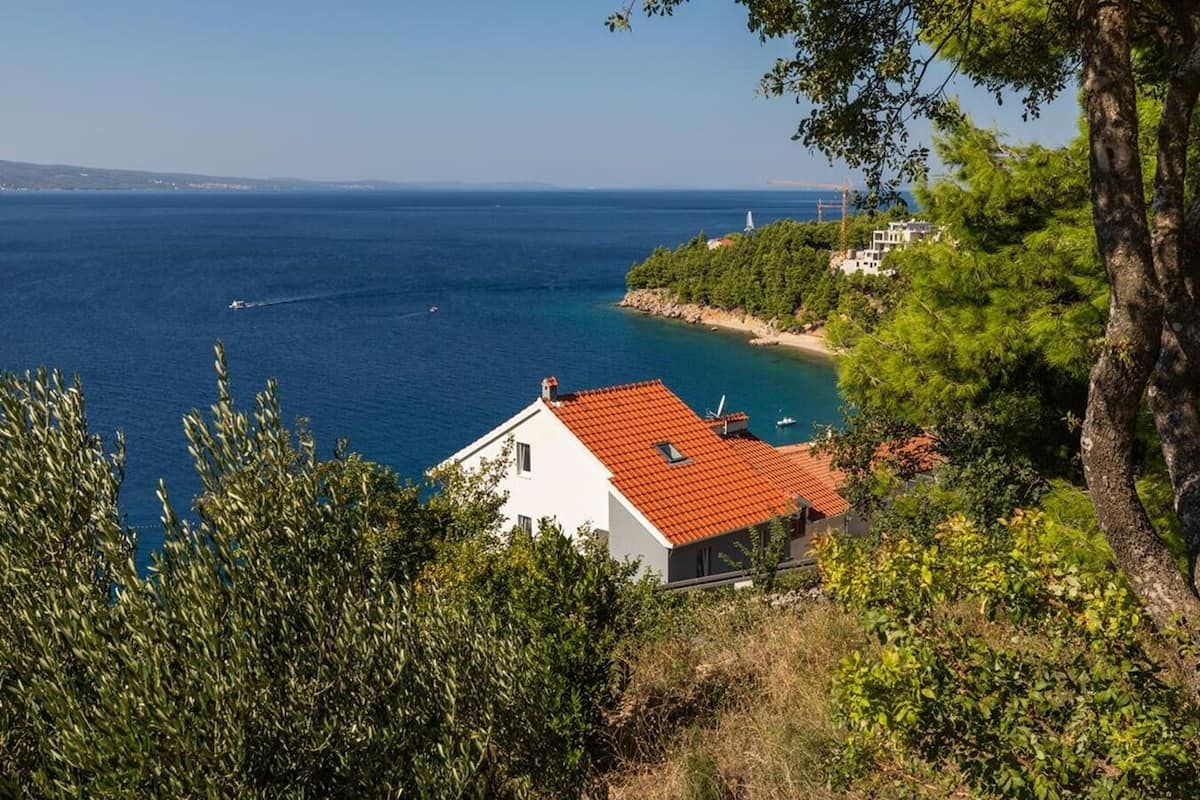 Una casa costera con techo de tejas rojas, rodeada de exuberante vegetación y con vistas a un mar azul y transparente.