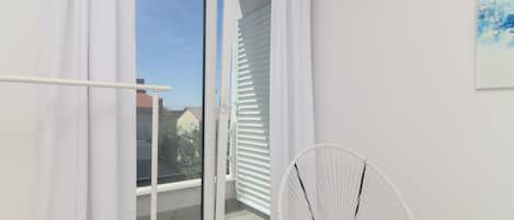 A balcony with white curtains, a white chair, and a clear view of houses in the background.