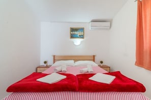 A neatly made bed with red and white bedding, a small bedside table with a plant, and a wall-mounted air conditioner. - House Seagull (Vir)