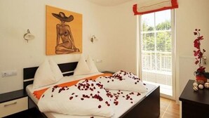 Een slaapkamer met een bed, een schilderij aan de muur, een raam met gordijnen en een nachtkastje met bloemen.
