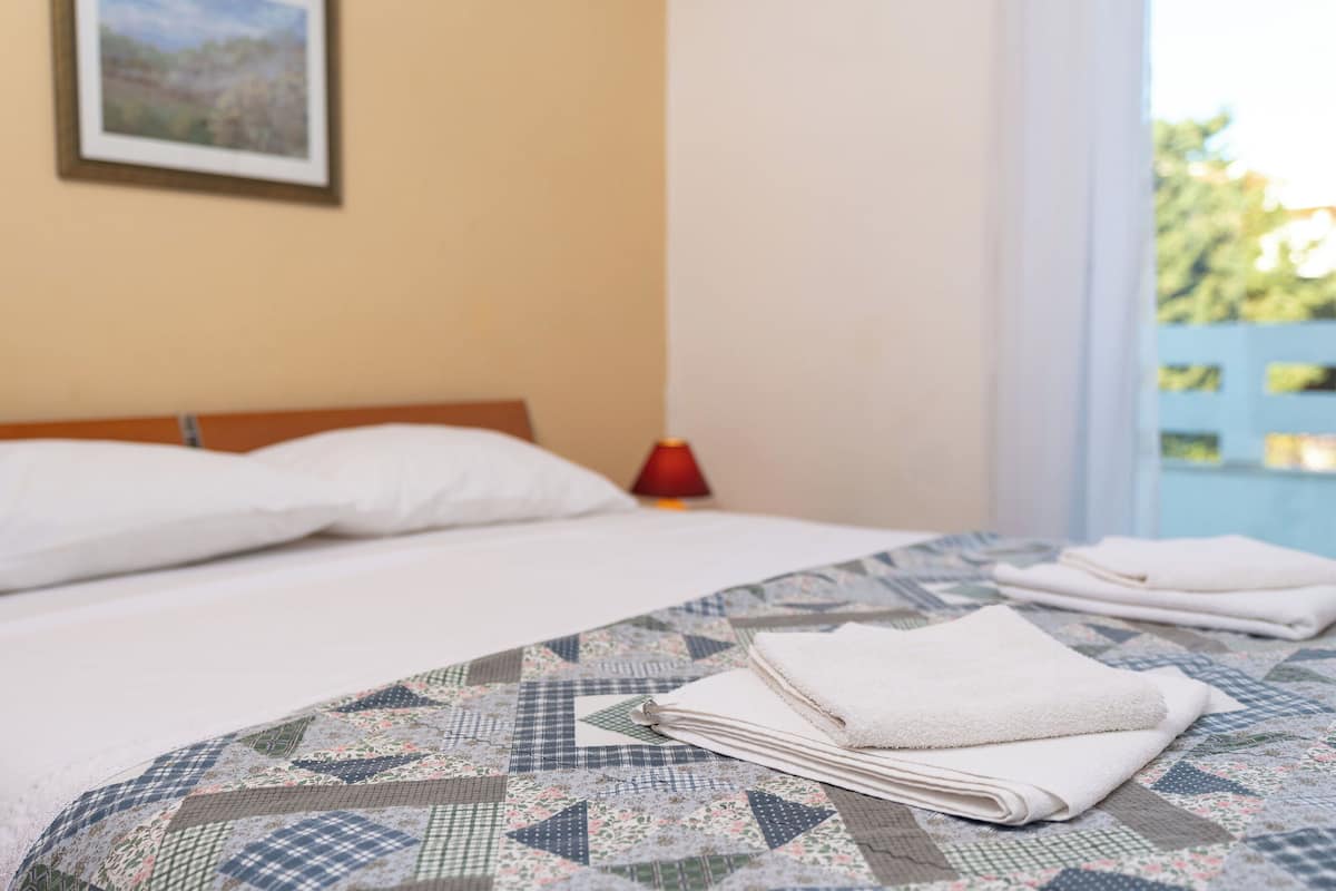 Una cama bien hecha con una colcha estampada, una pila de toallas blancas y una lámpara roja en la mesita de noche.