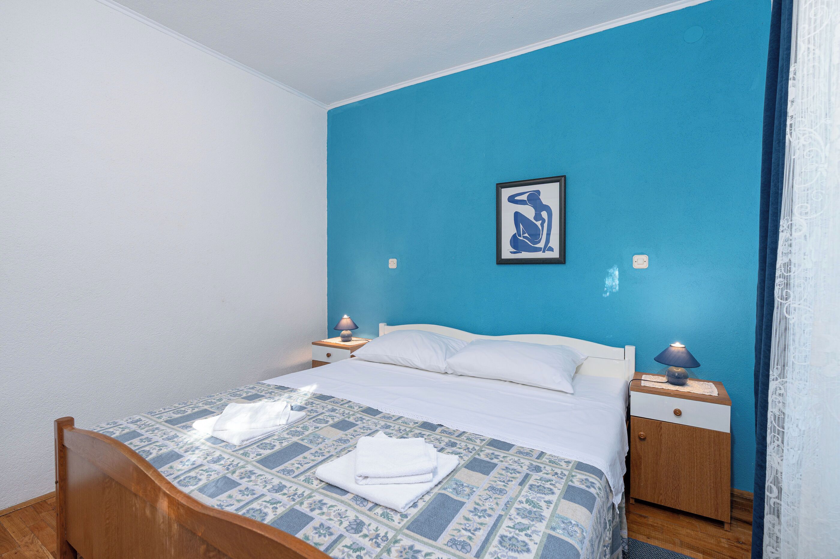 A bedroom with a bed, two bedside tables, a nightstand, a wall-mounted picture, and a window with curtains.