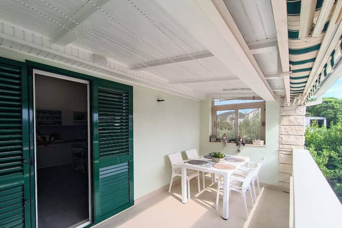 A balcony with a dining table and chairs, a green door, and a view of greenery.