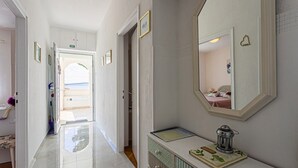 Un pasillo con un espejo, una mesita de noche con un libro y una vela, y una vista de un dormitorio reflejado en el espejo.