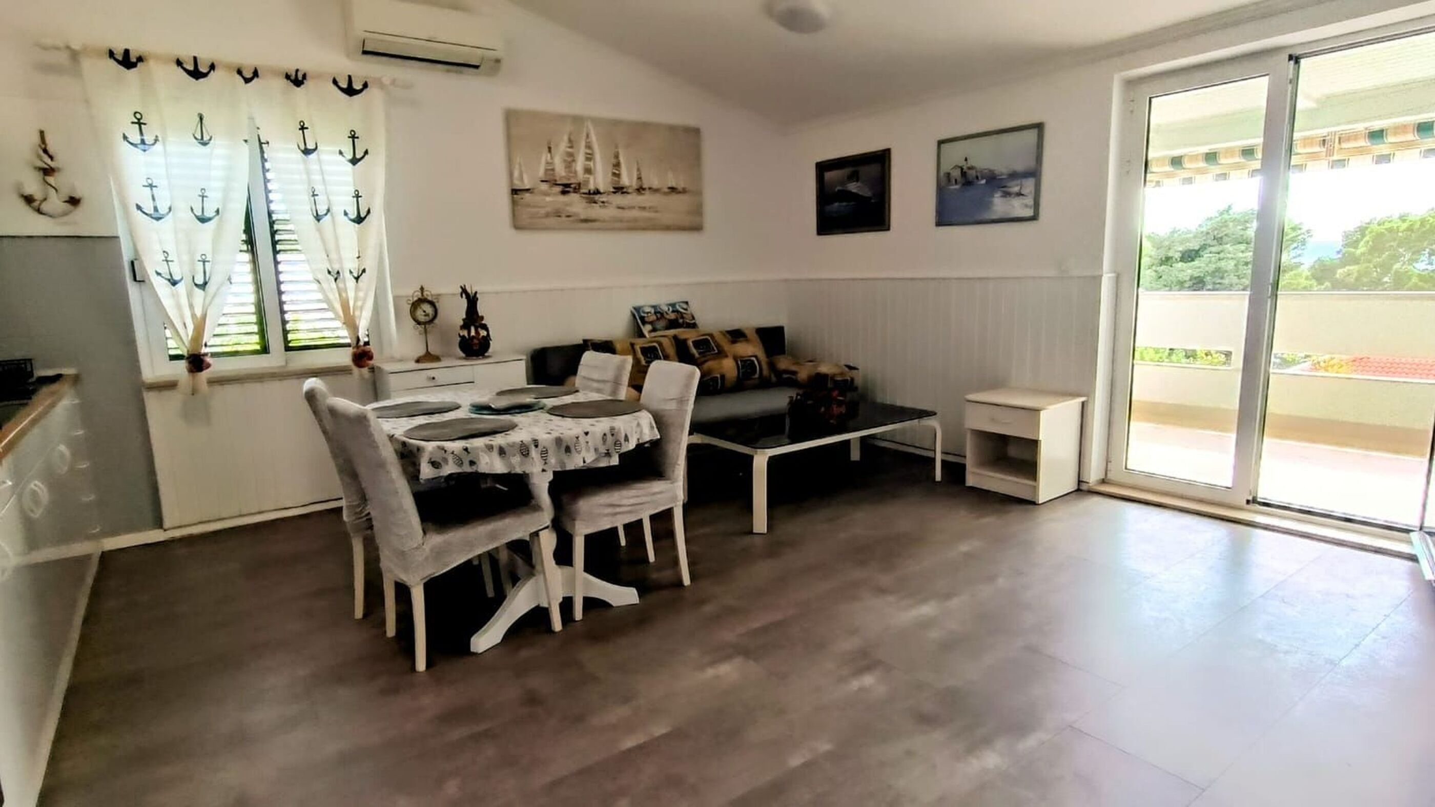 Et spiseområde med et bord og stole, en sofa og et lille hvidt sofabord. Der er to vinduer med gardiner, og billedkunst på væggene.