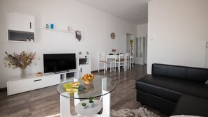 A modern living room with a flat-screen TV, a glass coffee table, a black leather sofa, and a dining area with white chairs and a table. - Apartment Miriam (Opatija)