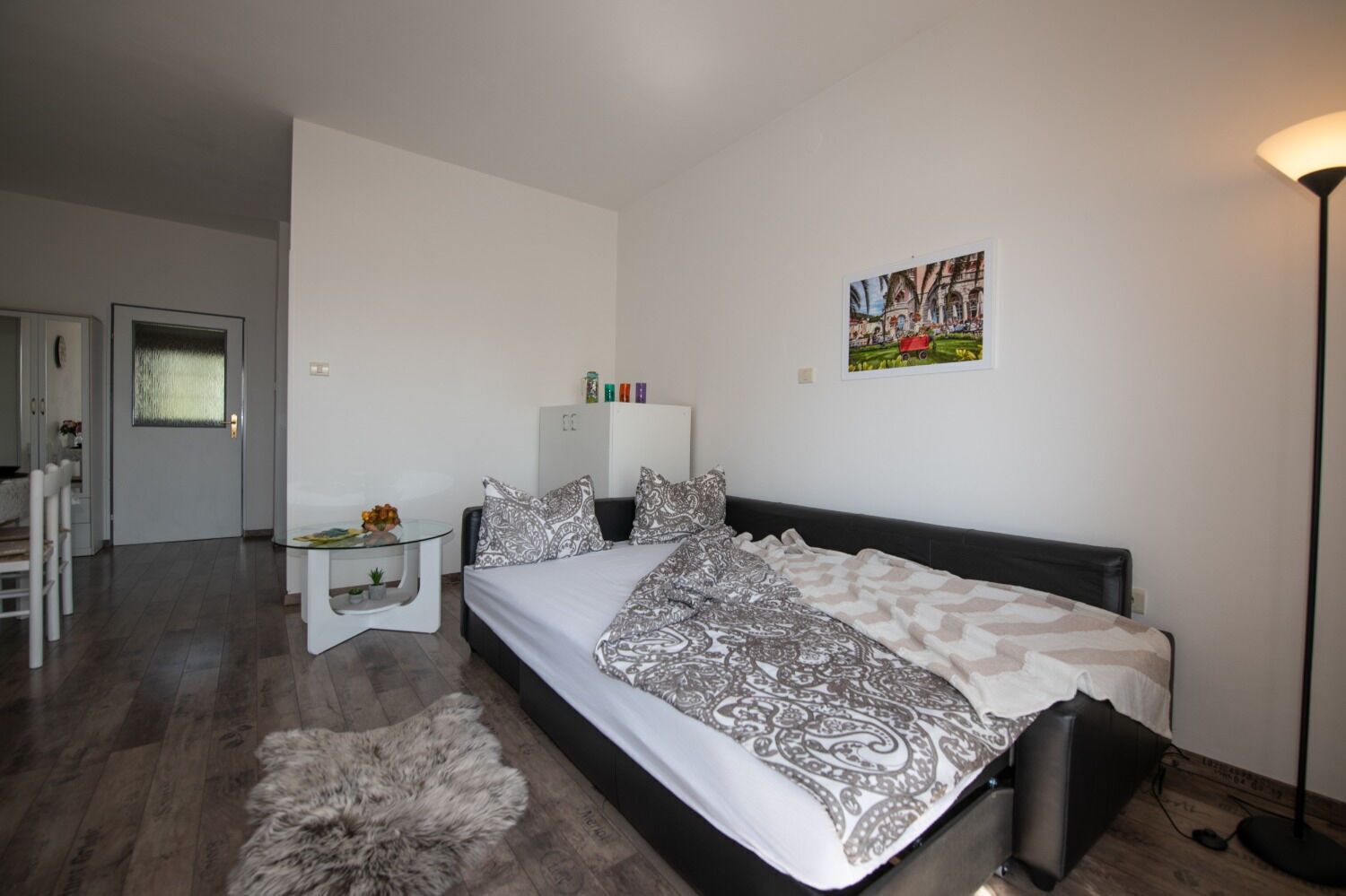 A bedroom with a bed, a glass table with a plant, a fur rug, a wardrobe, and a painting on the wall.