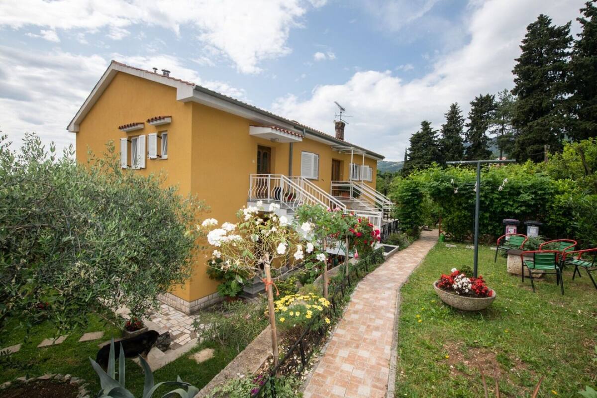 A yellow building with a staircase leading to a balcony, surrounded by a garden with various plants and flowers.