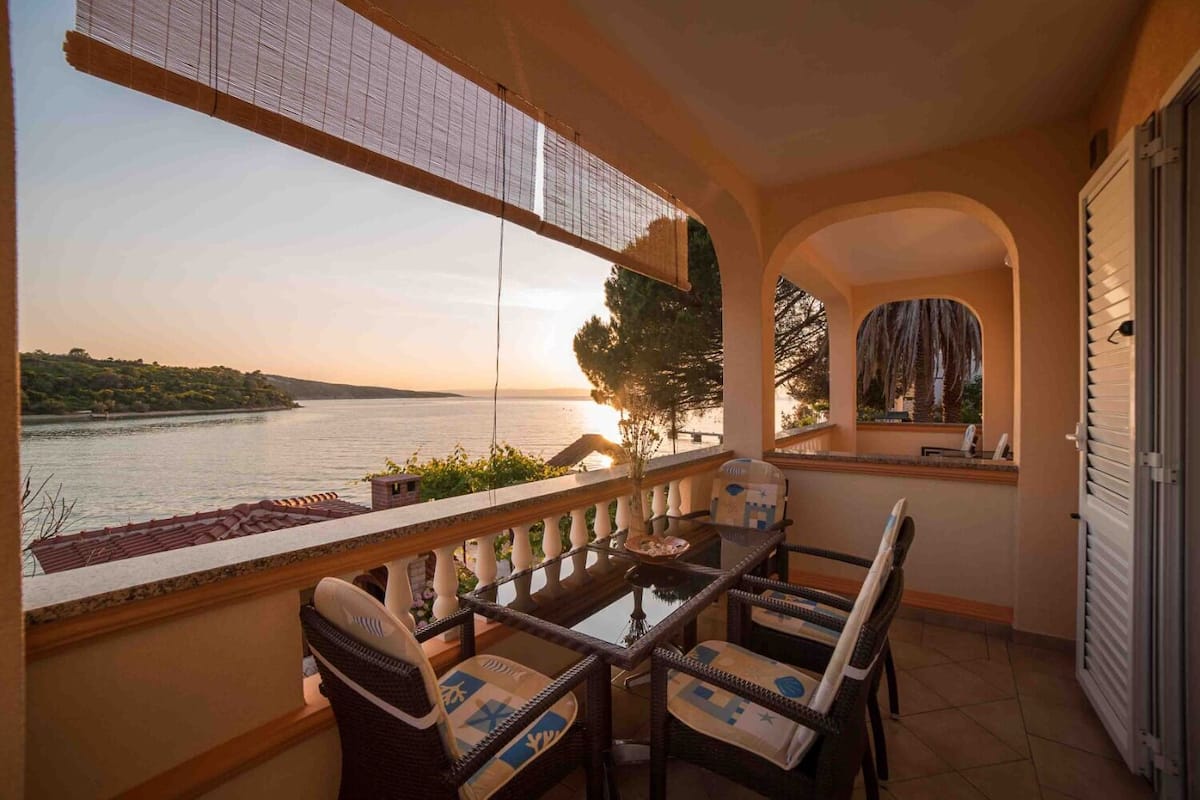 A balcony with a table and chairs, overlooking a body of water and a sunset.