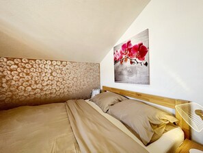 Makuuhuoneessa on sänky, kukka-aihekuvioitu pääty, yöpöytä lampun kanssa ja seinälle ripustettu kuva punaisista kukista.