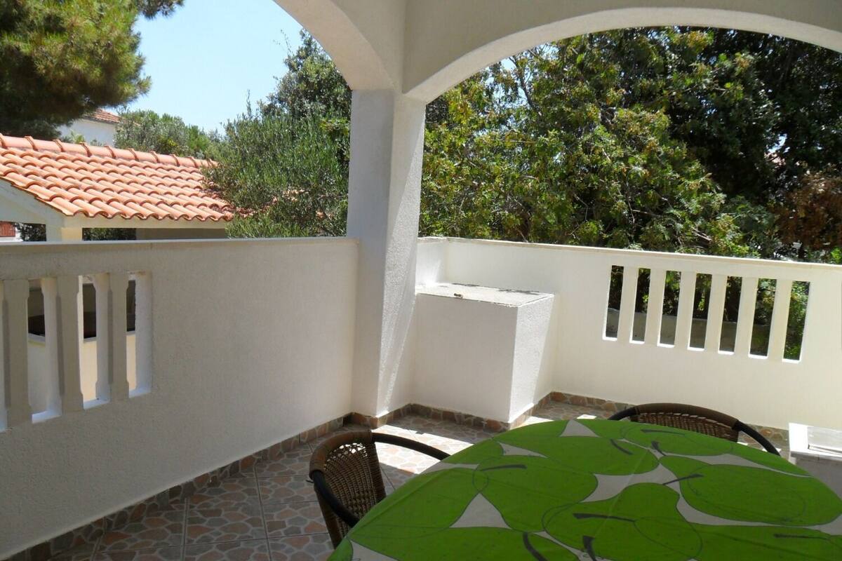 Un espace extérieur abrité, doté d’une table et de chaises, d’une rambarde blanche, et offrant une vue sur des arbres et un bâtiment dont le toit est recouvert de tuiles rouges.