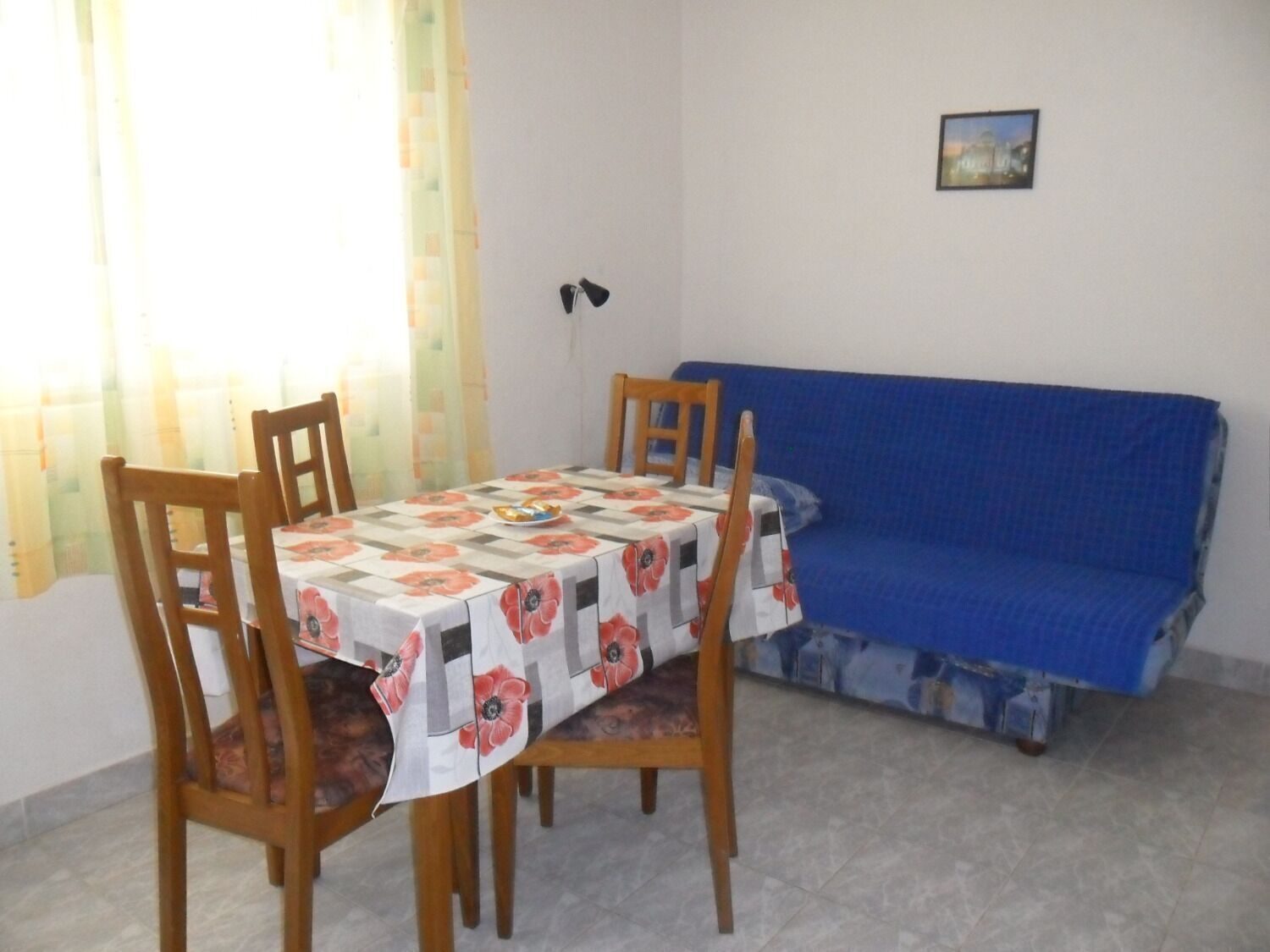 A dining area with a table covered by a floral tablecloth, surrounded by wooden chairs. A blue sofa is against the wall, and a small framed picture is on the wall above the sofa.