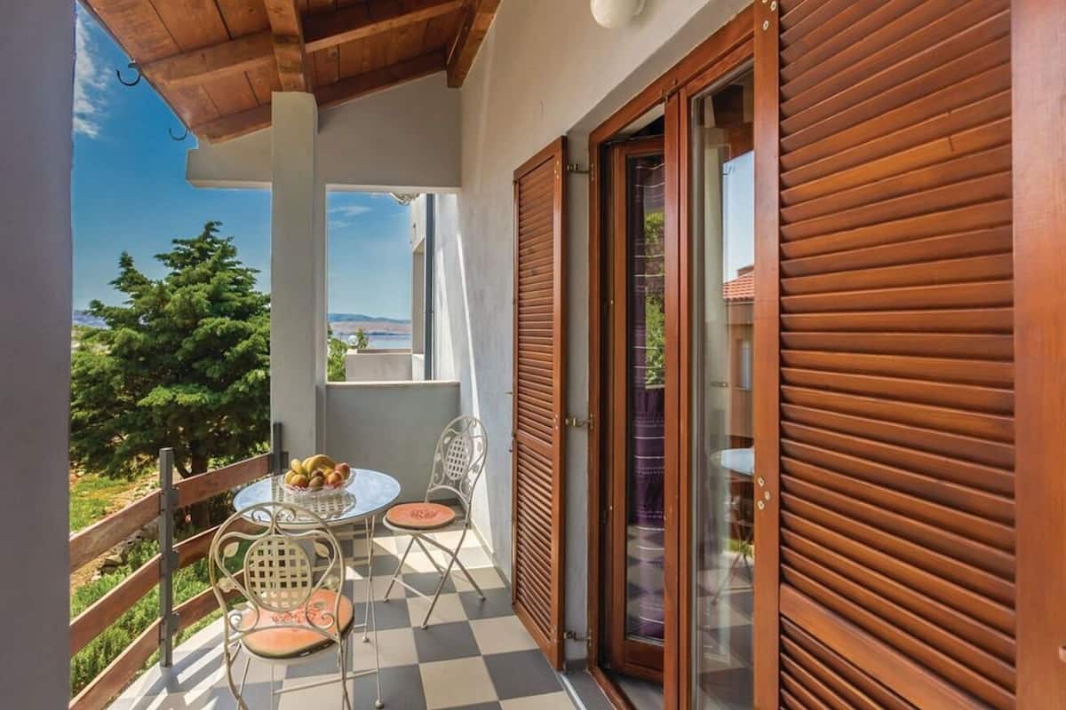 A balcony with a table and chairs, a view of trees and a distant landscape, and a wooden door with shutters.