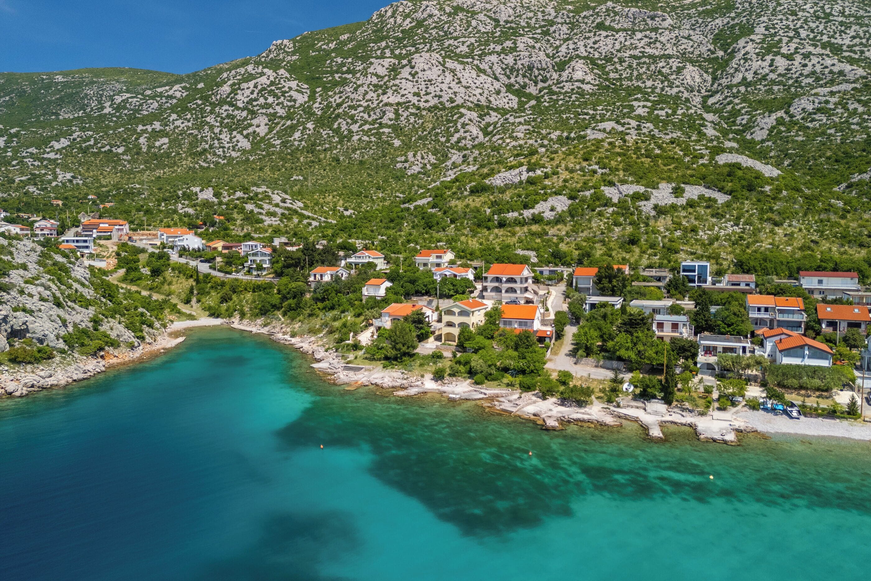 Vue aérienne d’un village côtier, avec des maisons le long du rivage, une mer turquoise d’un bleu limpide et un paysage montagneux en arrière-plan.