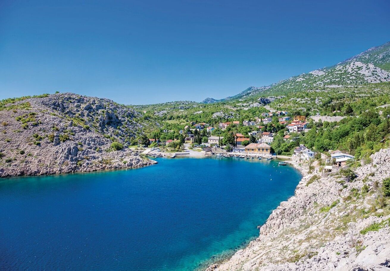 A coastal town with a clear blue sea, rocky terrain, and buildings with red roofs.