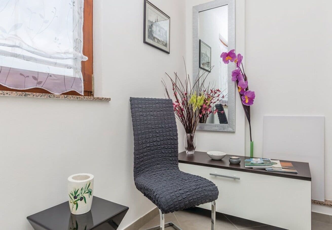 Een moderne kamer met een donkere, gestructureerde stoel, een klein bijzettafeltje met een decoratieve mok, een spiegel en een vaas met bloemen.