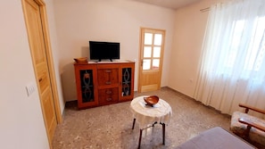 A room with a television on a wooden cabinet, a small round table with a bowl, a window with curtains, and a door. - Apartment Nadi (Mali Losinj)