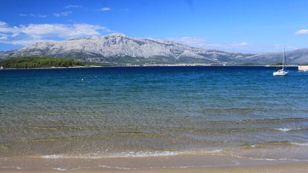 Ein Strand mit klarem Wasser, einer Segelyacht und einem Berg im Hintergrund.