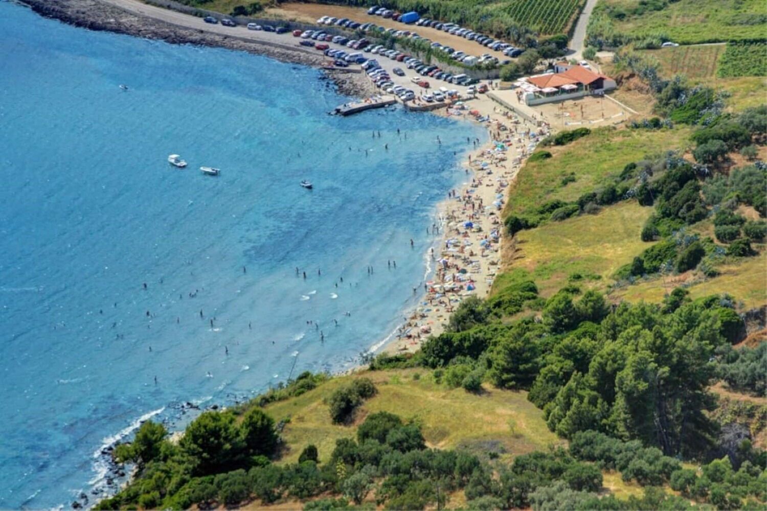 An aerial view of a coastal area with a beach, parked cars, and a building with a red roof.