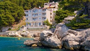 Um edifício de vários andares com varandas, localizado numa costa rochosa com águas azul-claras.