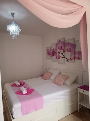 A bedroom with a bed, pillows, a bedside table, a chandelier, and orchid wall art. - Apartment Mira (Rab)