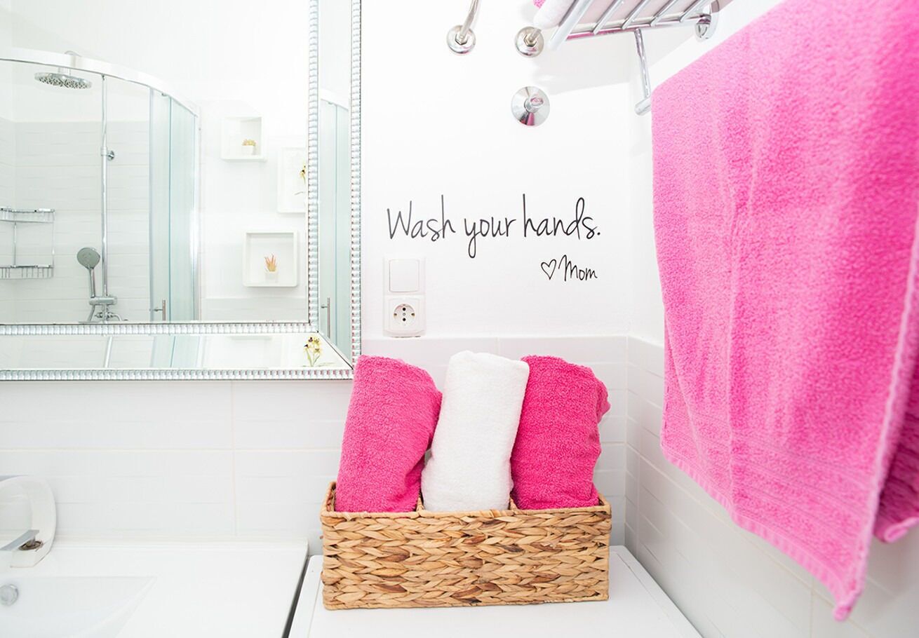 A bathroom with a glass shower enclosure, a white sink, a mirror, a wall-mounted towel rack with a pink towel, and a basket with pink and white towels.