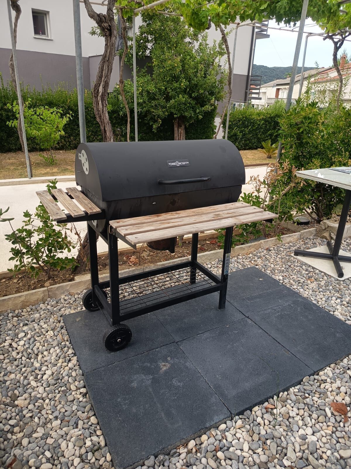Una parrilla portátil para exteriores con un banco de madera sobre ruedas, ubicada sobre una superficie de grava con una zona pavimentada cerca.