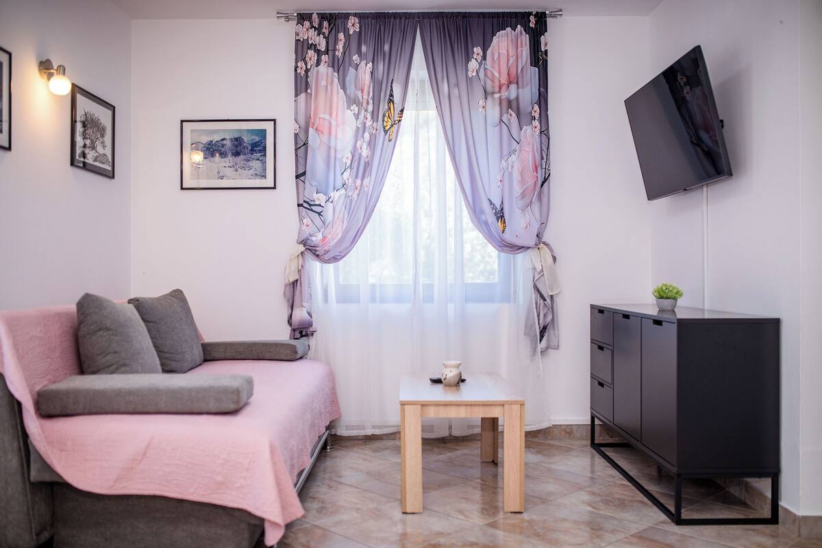 A bedroom with a bed, a small table, a TV mounted on the wall, and a window with curtains.