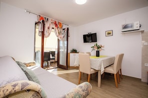A modern living room with a sofa, a dining table with chairs, a TV mounted on the wall, and a sliding glass door leading to an outdoor area. - Apartments Zdenka (Rab)