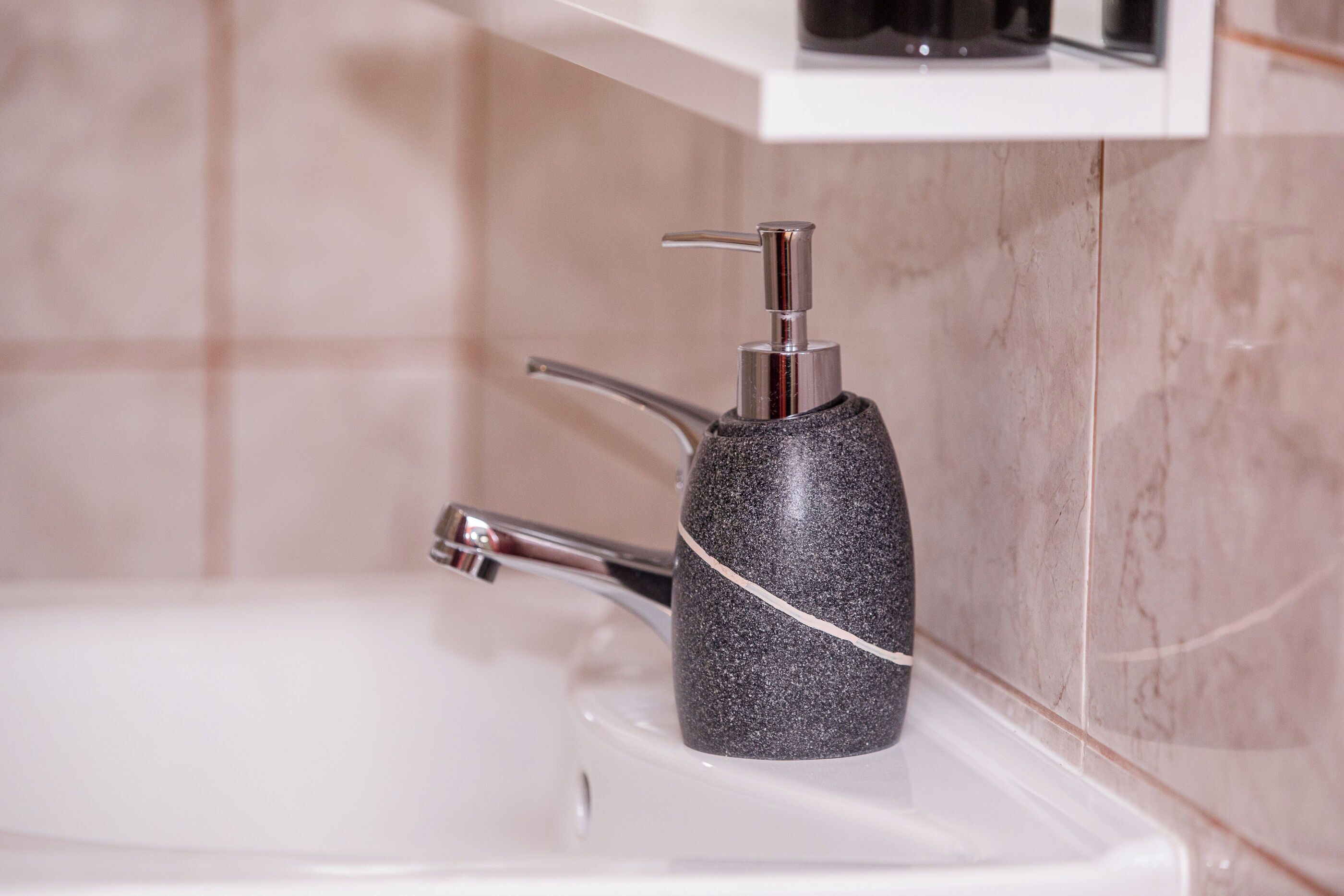 Une salle de bain avec un mur en marbre, un lavabo blanc et un distributeur de savon noir.
