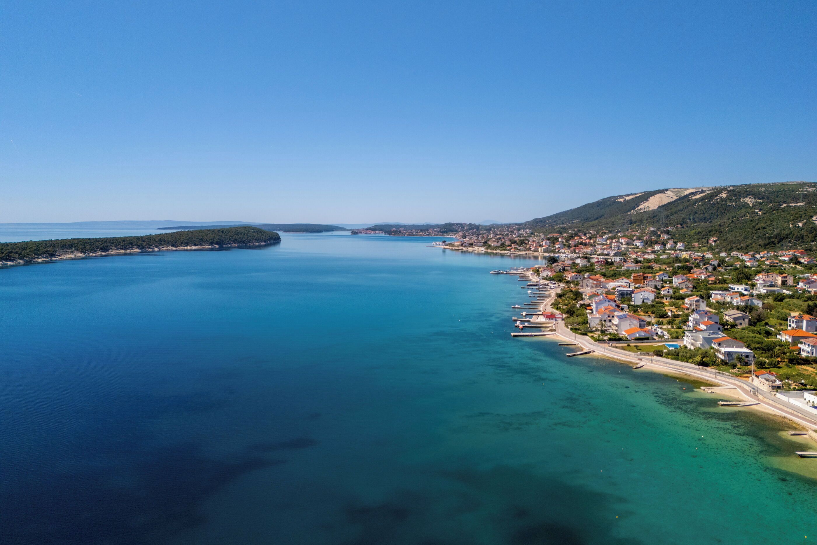 Une ville côtière dotée d’un port de plaisance, d’eaux d’un bleu limpide et d’un paysage vallonné en arrière-plan.