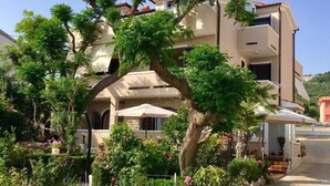 A multi-story building with a large tree in the front yard, surrounded by various plants and flowers. - Apartments Zdenka (Rab)