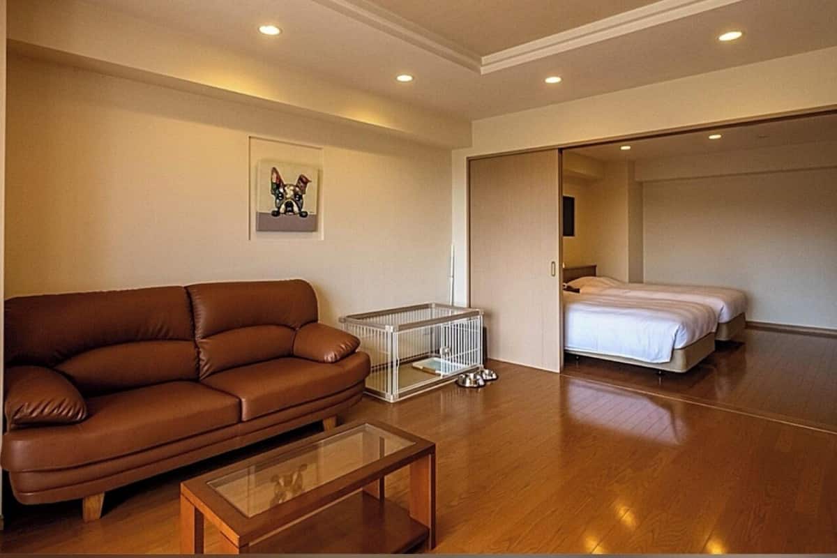 A living room with a brown leather sofa, a glass coffee table, a white pet cage, and a bed in the adjacent room.