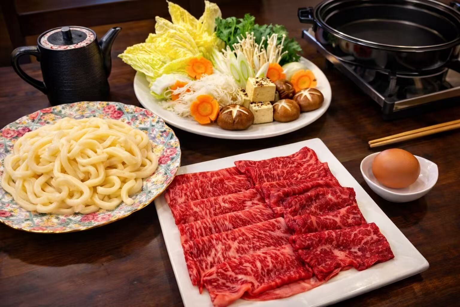 A table with a plate of noodles, a plate of sliced beef, a bowl of egg, and a teapot.