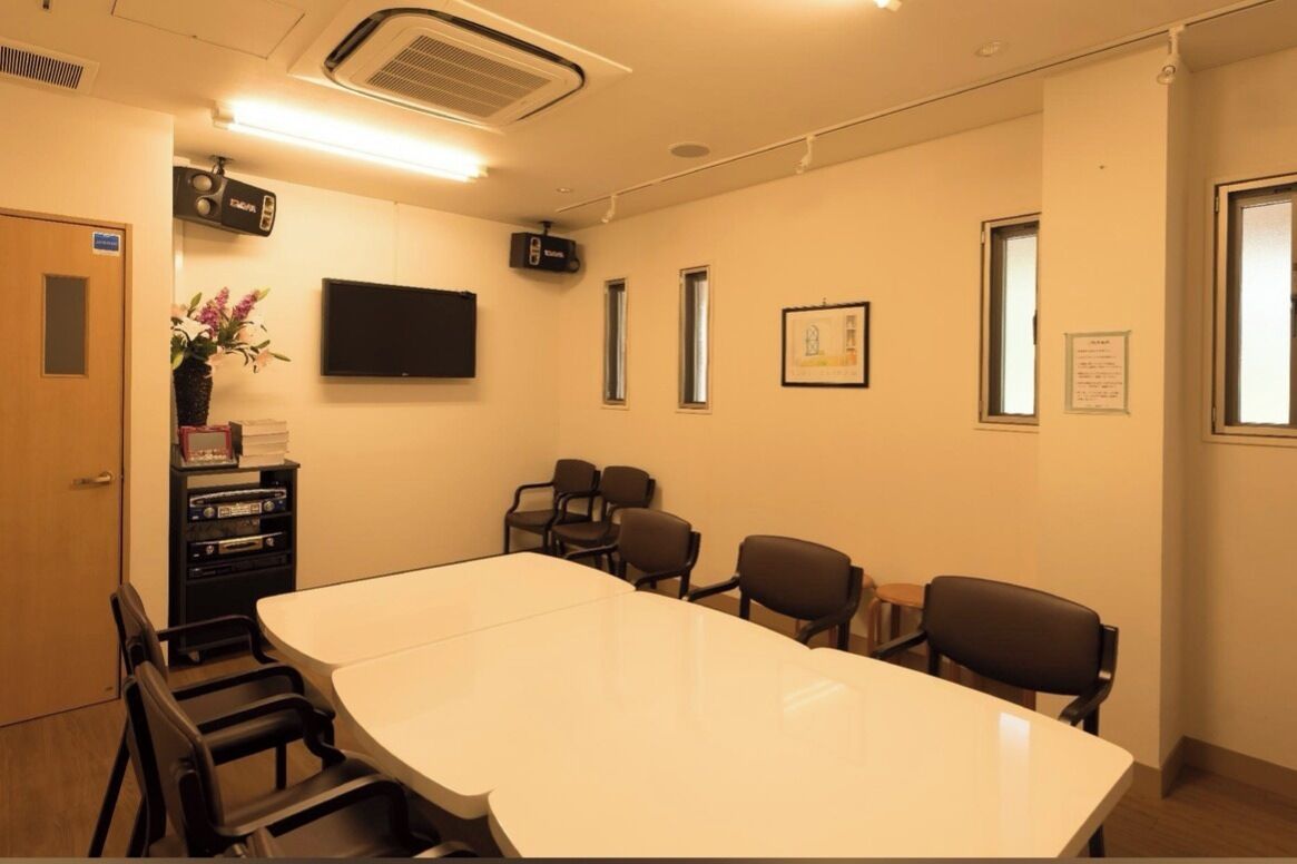 Un salón de conferencias con una gran mesa, varias sillas, un televisor de pantalla plana, un proyector y una pequeña planta en un estante.
