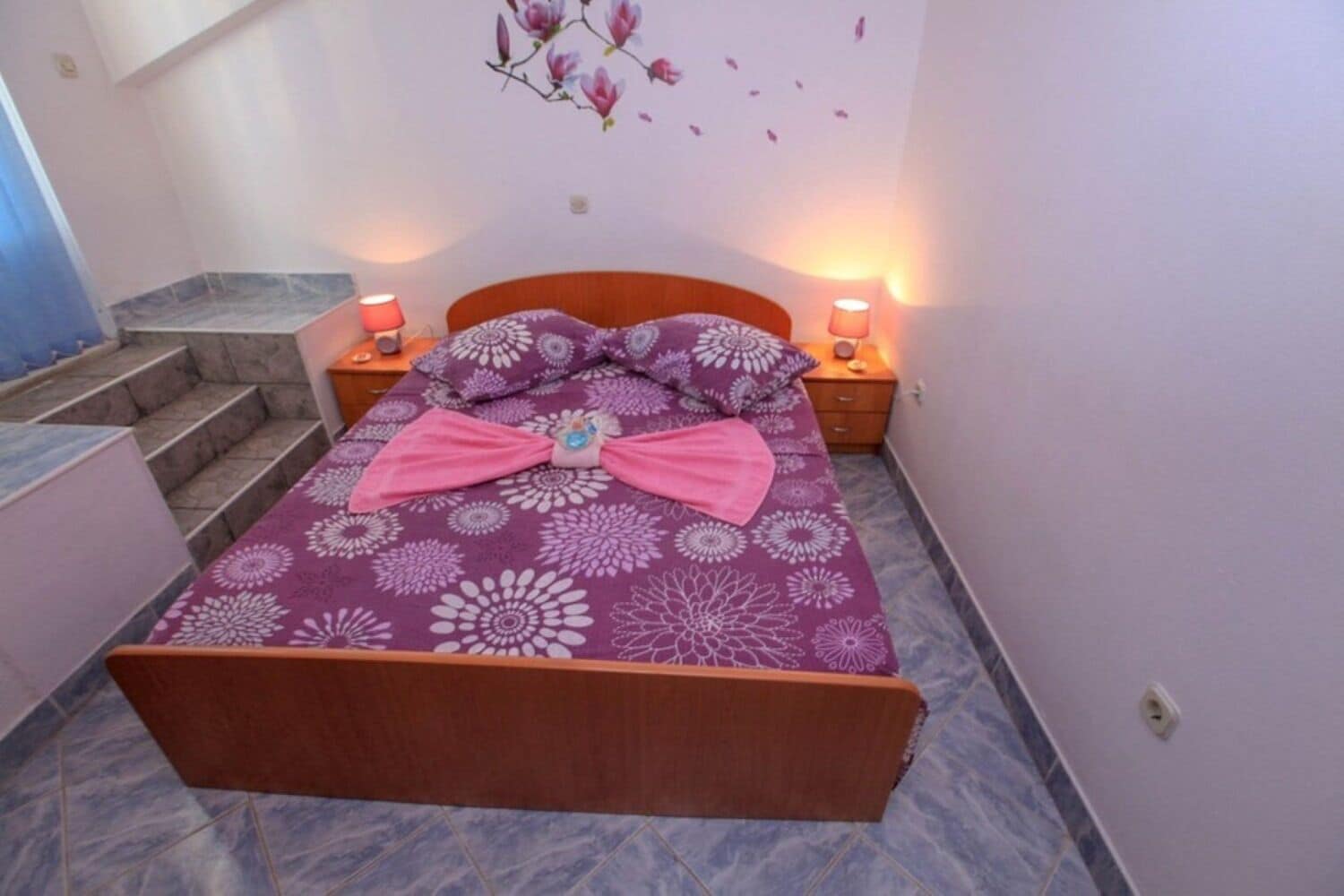 A bedroom with a bed, two bedside tables, a nightstand, a wall-mounted lamp, and a staircase.