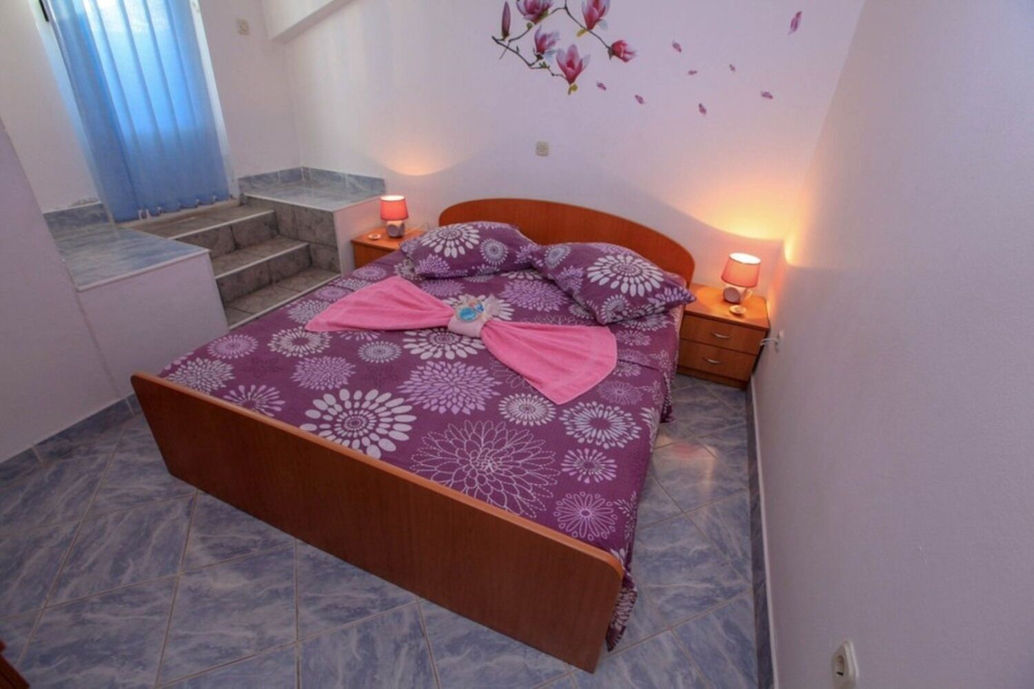 A bedroom with a bed, two bedside tables, a nightstand, a window with curtains, and a wall-mounted flower decoration.