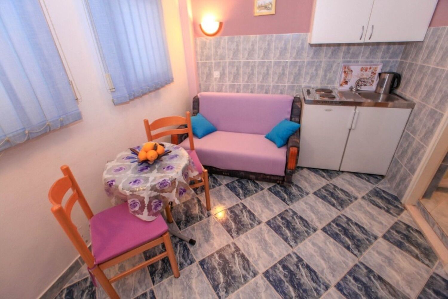 A compact kitchen with a purple sofa, a small dining table with a floral tablecloth, and a fruit bowl.